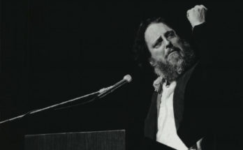 A photo of Peter Schickele speaking at a podium and clenching his fist.