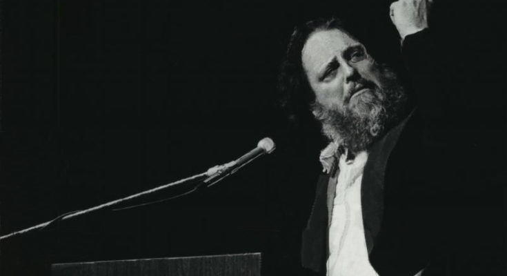 A photo of Peter Schickele speaking at a podium and clenching his fist.